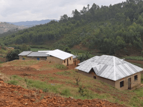 Casas simples com telhado metálico, solo avermelhado e montanha com vegetação ao fundo